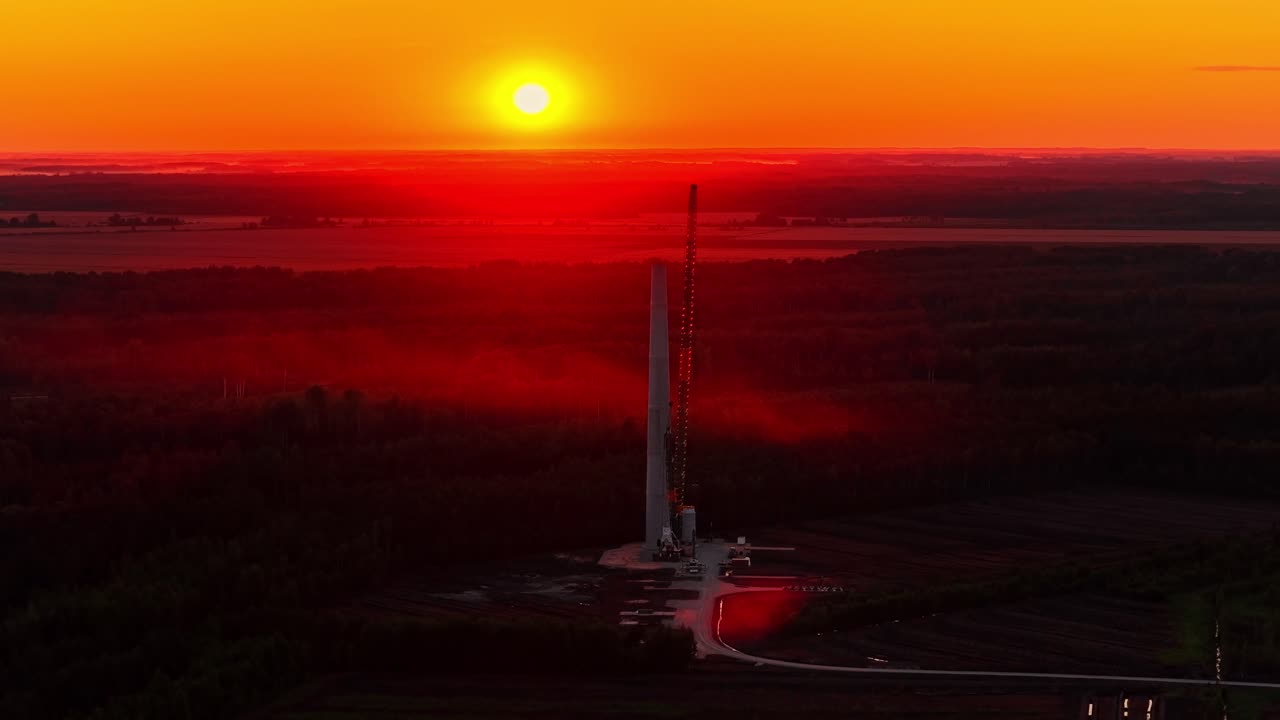 Slow aerial drone shot approaches a huge wind turbine construction site with a large crane, silhouetted against a beautiful orange sunset in the Latvian countryside