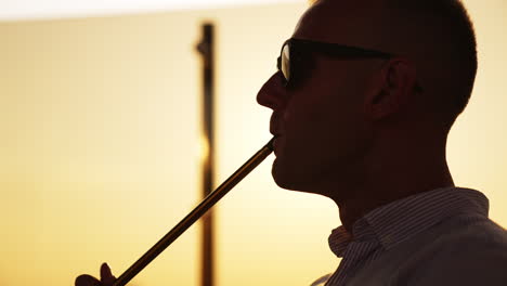 upper part of a man in sunglasses breathing in and out thick white smoke. young man smoking hookah at sunset close up.