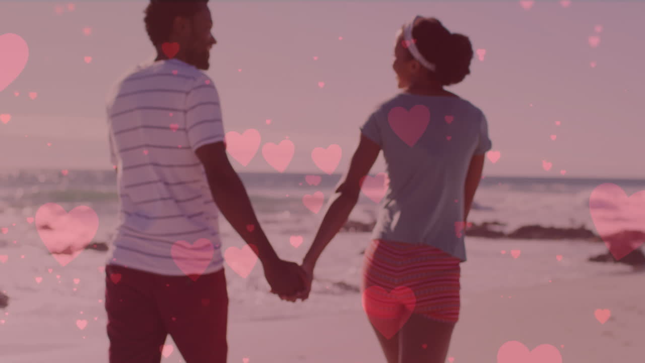 아프리카계 미국인 커플이 손을 잡고 해변에서  ⁇ 고 있을 때 여러 개의 분홍색 심장이 떠다니고 있습니다.