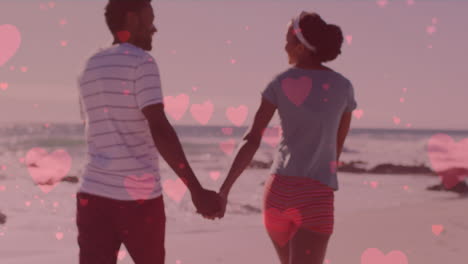 multiple pink hearts floating against african american couple holding hands and walking on the beach