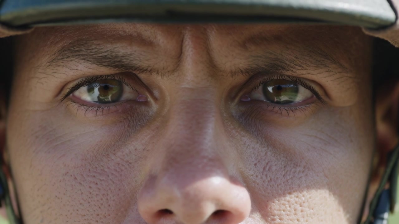 Camera centering soldier holding steady gaze watching operator in sun, with helmet reflection