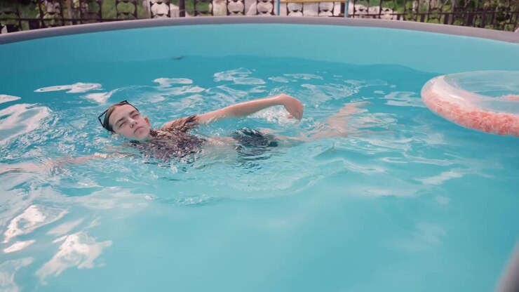 Young woman swims in bright blue pool performing various strokes and floating on her back. scene includes clear water with gentle ripples inflatable ring float nearby and outdoor