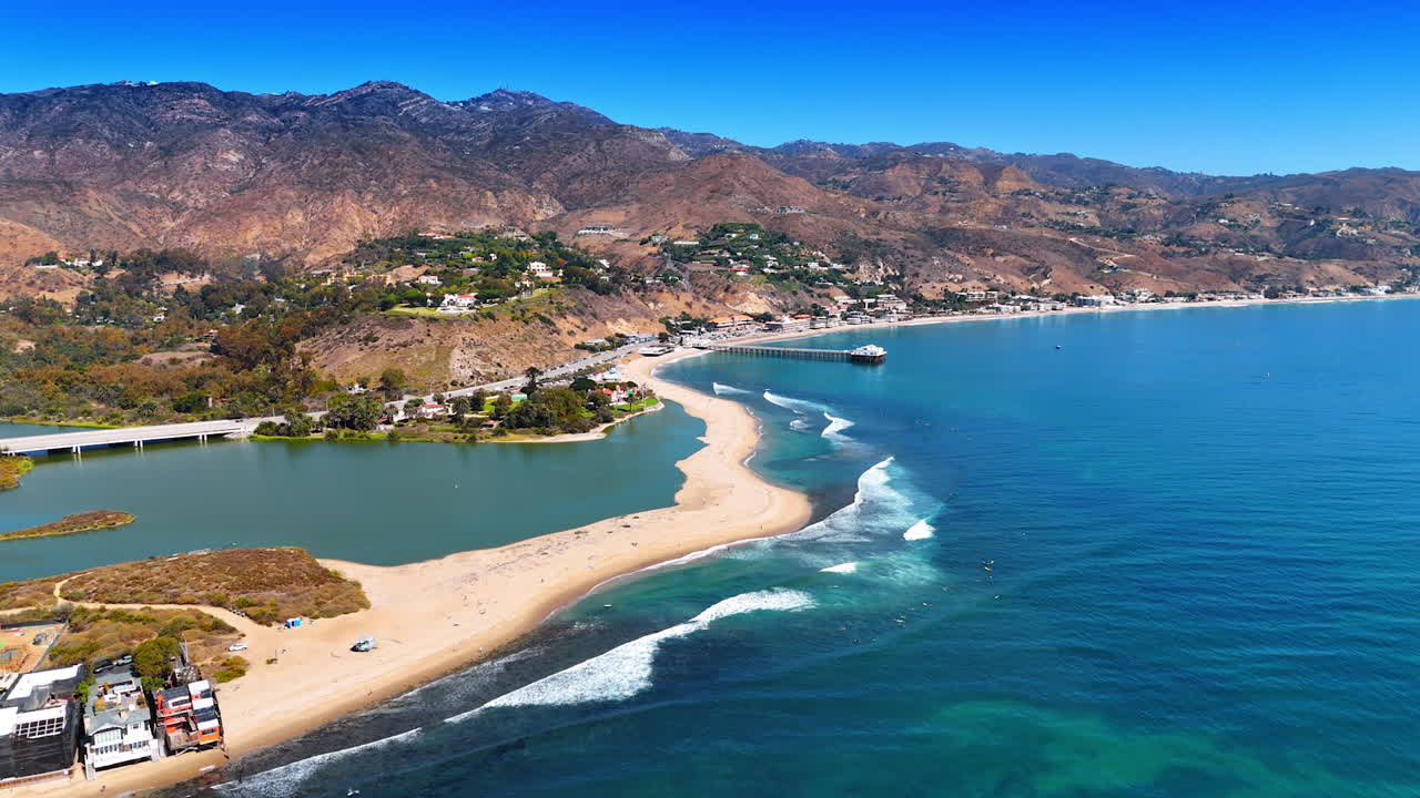 Approaching the narrow line of sandy beach meeting foamy waves. Green residential area scattered on the slopes of the rocks. Malibu, California, USA. Aerial view