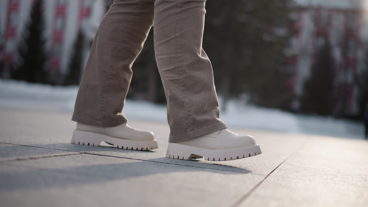 Leg view of choreographer tapping and dancing through snowy park plaza, lost in fluid choreography, wearing chunky white platform shoes and beige corduroy pants on tiled ground with long pale shadows