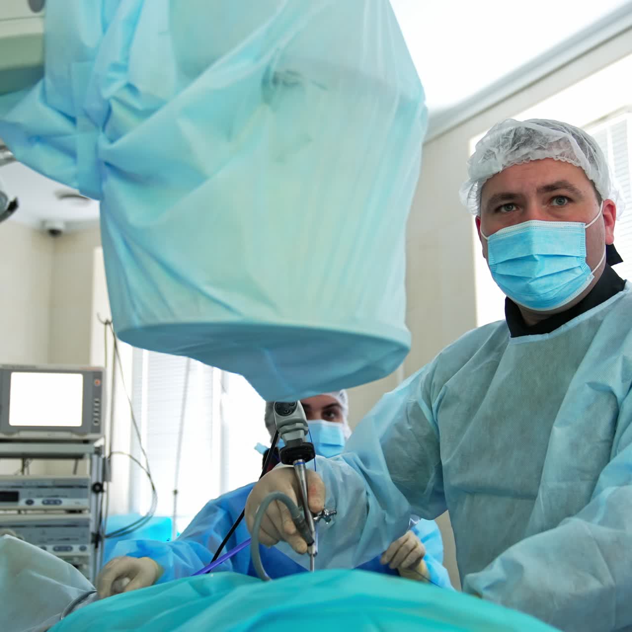 Adult doctor uses the tool during the operation. Surgeon looks attentively at the screen in front of him. Equipped surgery room at backdrop