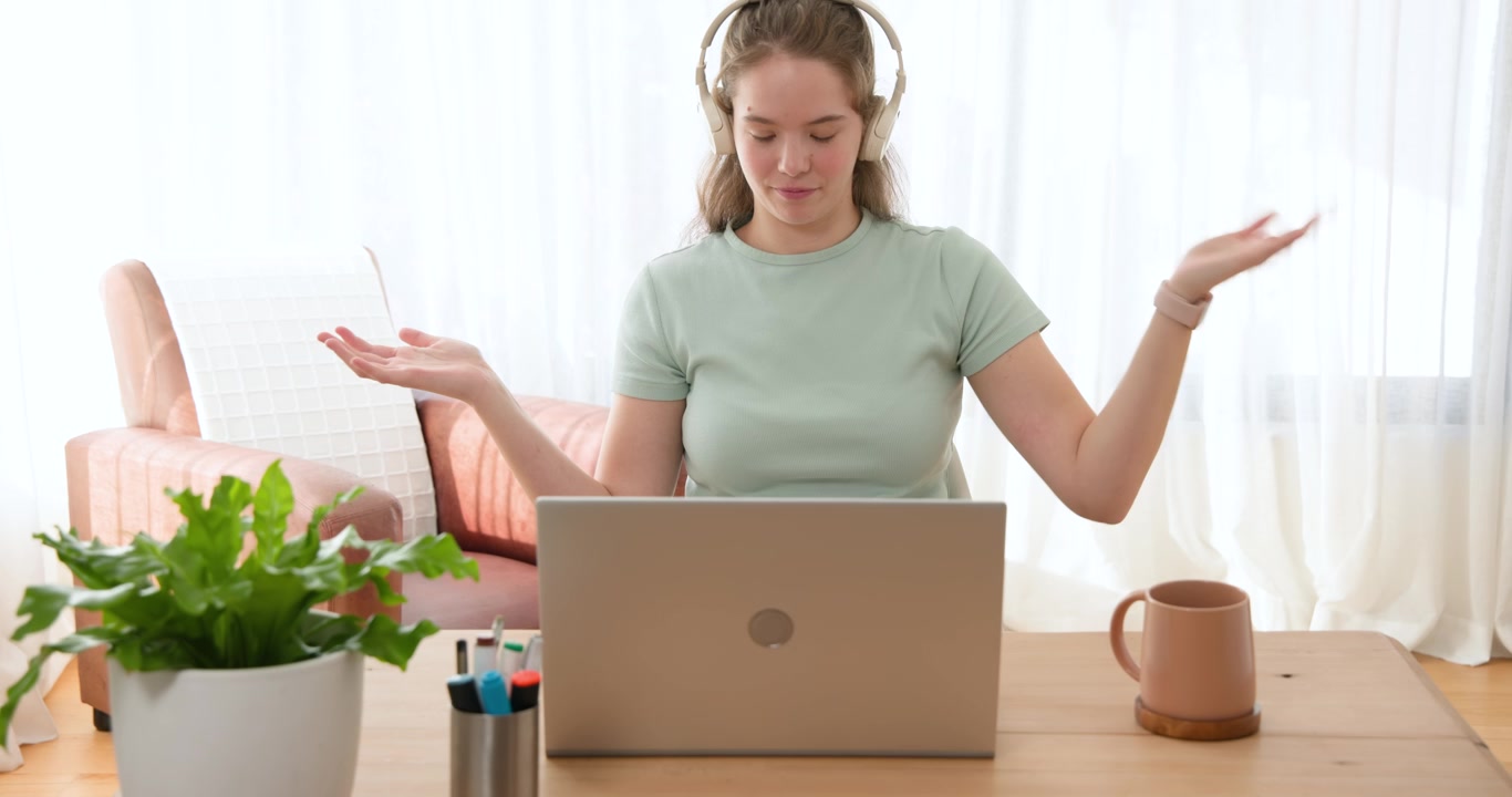 Frau in grünem T-Shirt mit weißen Kopfhörern gestikuliert am Laptop und reagiert auf einen Besprechungshinweis am Schreibtisch.