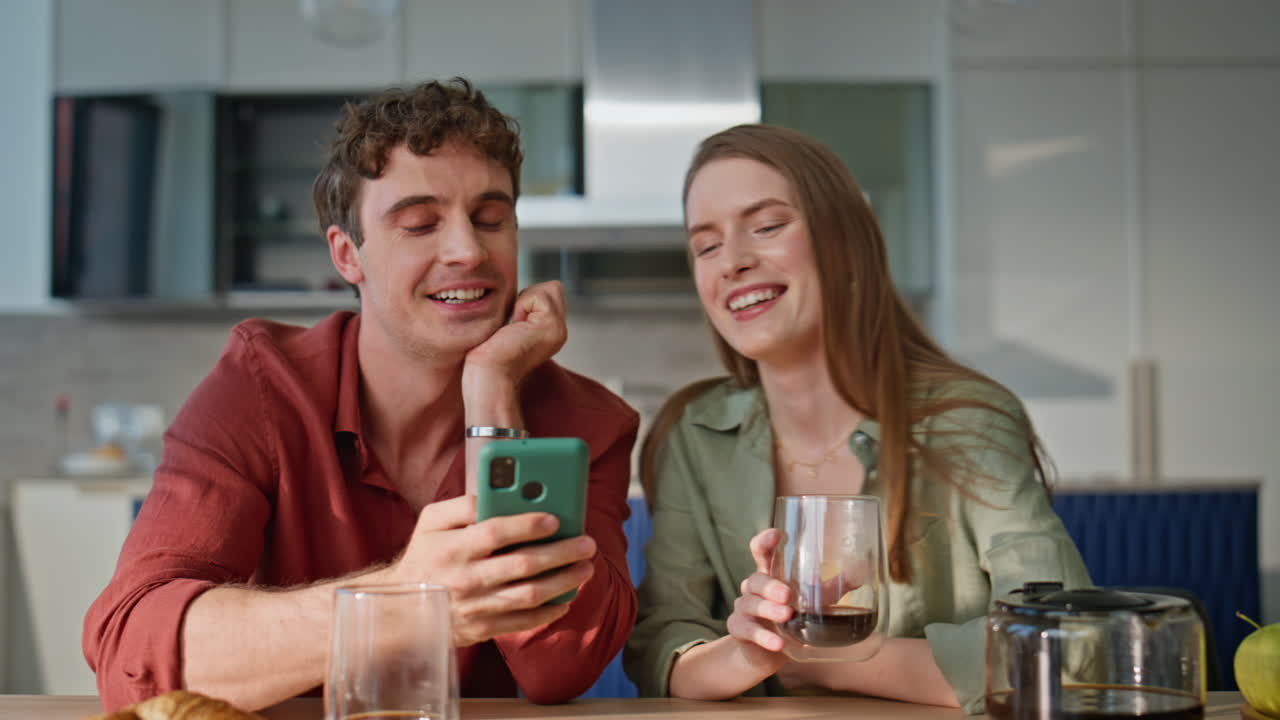 Laughing couple looking cellphone sitting at home kitchen closeup. Happy spouses