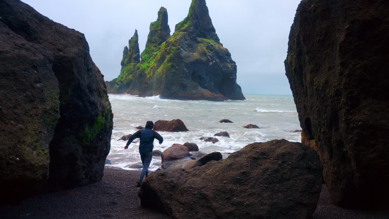 hombre corriendo a través de rocas en reynisfjara playa de arena negra en islandia con vistas a la formación de roca basáltica reynisdrangar en segundo plano.