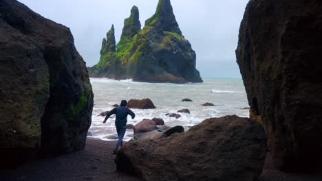 hombre corriendo a través de rocas en reynisfjara playa de arena negra en islandia con vistas a la formación de roca basáltica reynisdrangar en segundo plano.