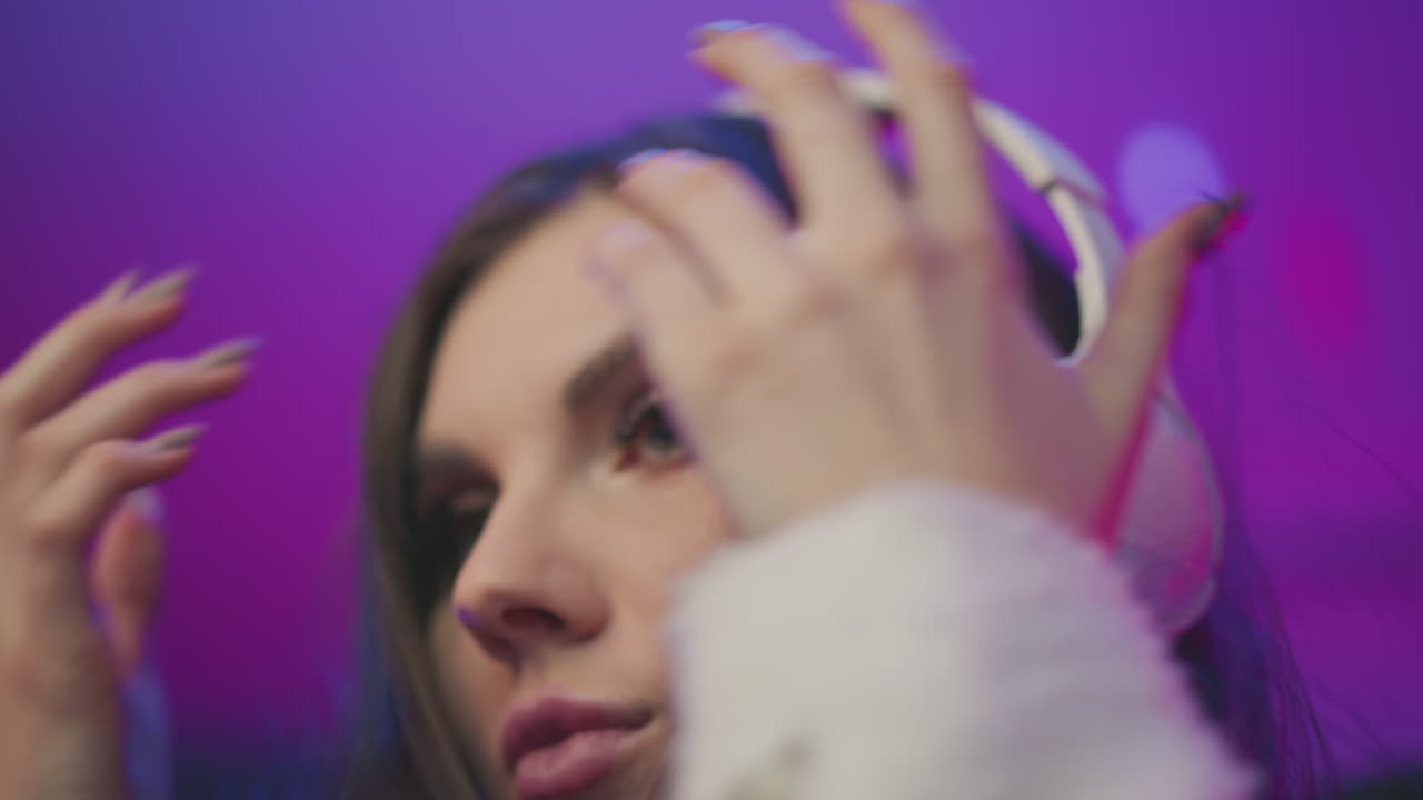 Close up lady raising white headphones toward her head in soft colorful ambient room with purple background, eyes closed, calm expression, cozy sweater sleeve visible