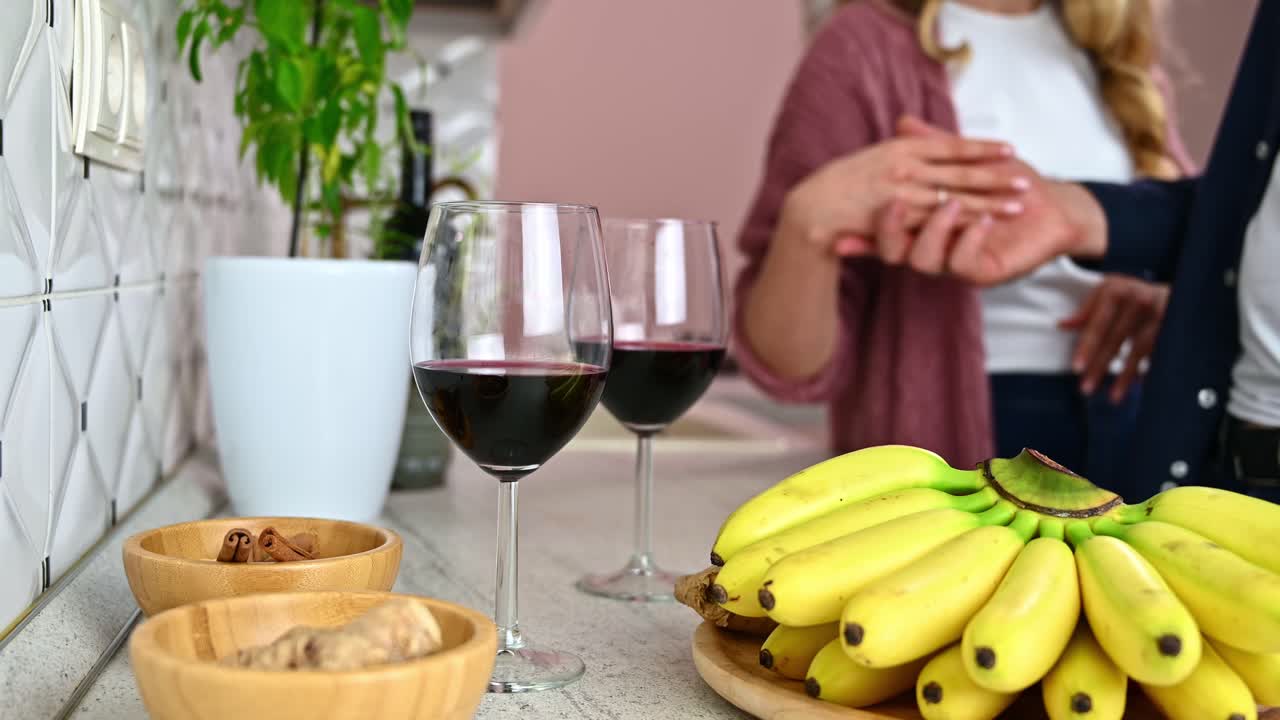 Man's hands lovely caress an woman hands in a white modern kitchen on blurred background. Glasses with red wine, bananas, cinnamon and ginger on wooden tables in foreground. Valentines day in love
