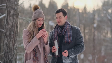 Schönes-Und-Stilvolles-Hipster-Paar-Aus-Mann-Und-Frau-In-Mantel-Und-Schal-Trinken-Nach-Einem-Spaziergang-Tee-Aus-Einer-Thermoskanne-Im-Winterwald.-Liebesgeschichte