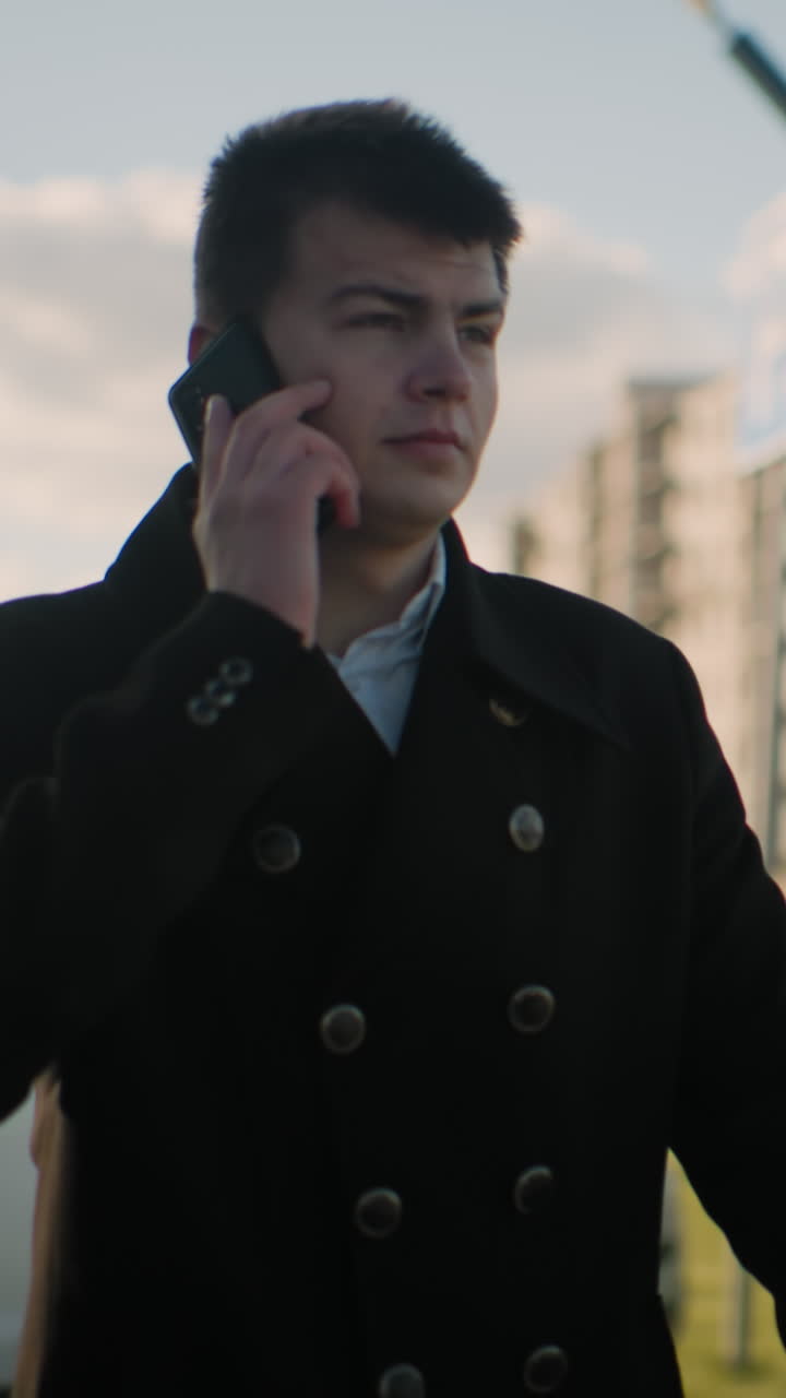 검은 코트를 입은 투자자는 주머니에서 전화를 꺼내서 심각한 표정으로 차를 떠나는 동안 주차된 자동차, 현대적인 건물, 움직이는 차량 및 초록색을 특징으로하는 도시 지역에서 전화에 응답합니다.