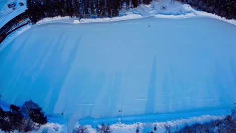 aerial rise over massive outdoor skating rink with less than a handful of people out during a twilight sunset with shadow reflections of tall pointy pine trees by public parks cabana onto the left1-2