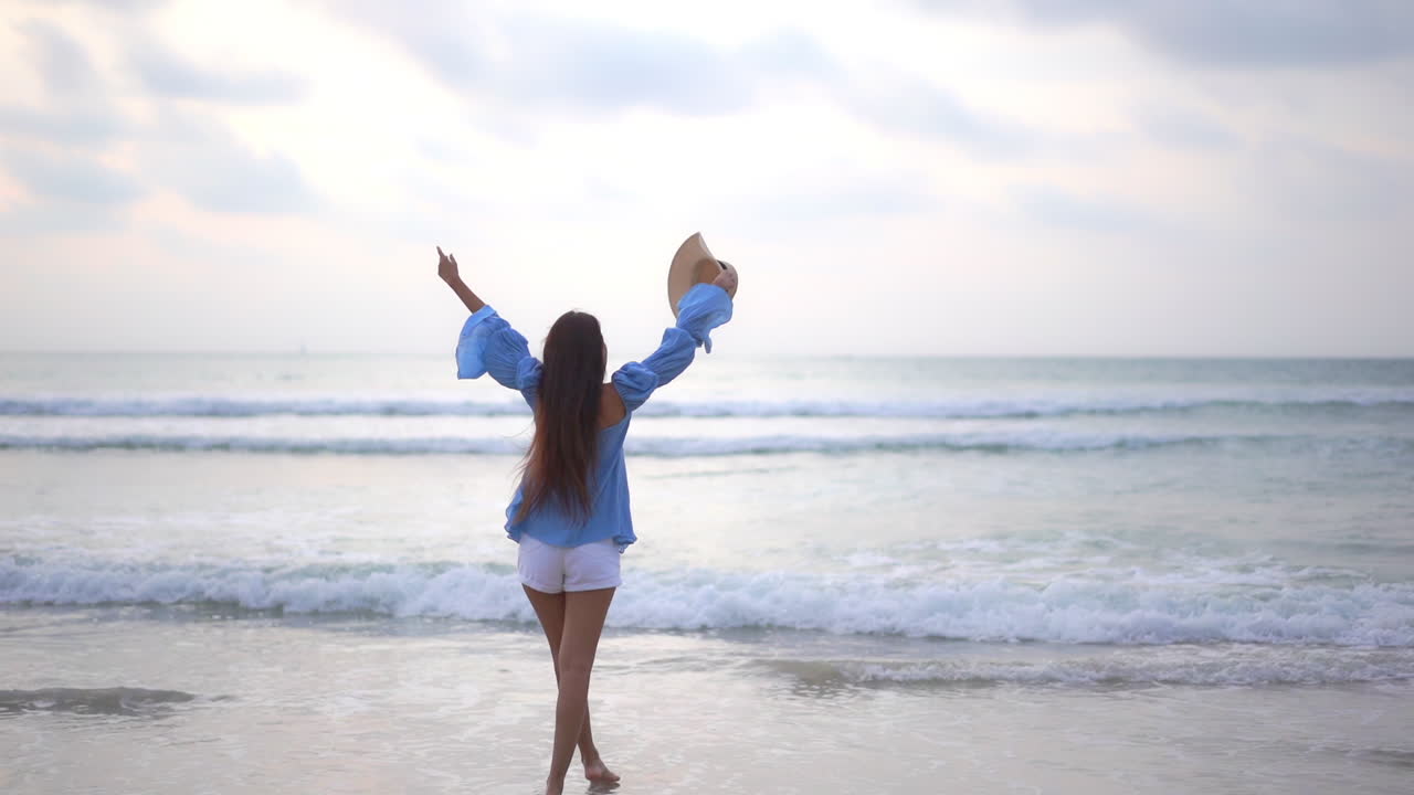 해변에서 바다를 향해 걸어가는 아시아 여성이 뒤돌아 일몰 흐린 하늘에서 팔을 들어 올립니다 - 휴가 템플릿 백뷰