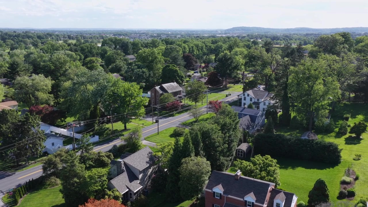 Suburban houses line tree-shaded street near Delp Road in Lancaster Pennsylvania, brick homes and manicured lawns spread beneath dense summer tree canopy and parked cars. Aerial drone flyover shot