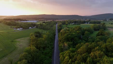car on rural intersection road during golden sunset in suburb. aerial descend wide shot. idyllic landscape with hilly agricultural farm fields and mountains in back. pennsylvania, usa.