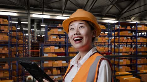 vista lateral de cerca de una ingeniera asiática con casco de seguridad mirando la tableta en su mano y mirando a su alrededor mientras está de pie en el almacén con estantes llenos de productos de entrega