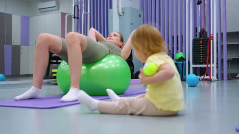 toddler holding tennis balls, one bouncing from hand, while adult rests on green exercise ball in gym, fitness environment includes purple mat, workout equipment, and modern decor