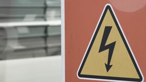 Blurred-tramway-running-behind-a-yellow-electrical-danger-sign-with-a-black-bolt-arrow