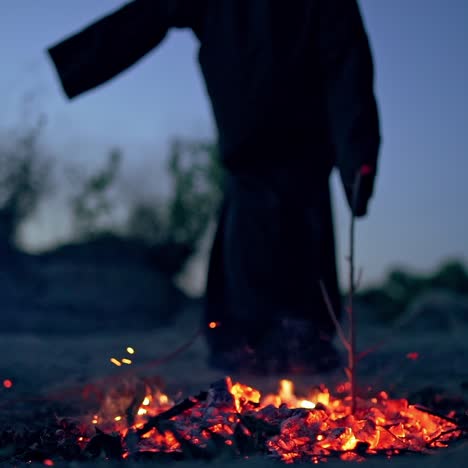 spooky death in black gown near fire. embers of fire on the ground and mysterious figure doing shamanism with a wooden stick. halloween day.