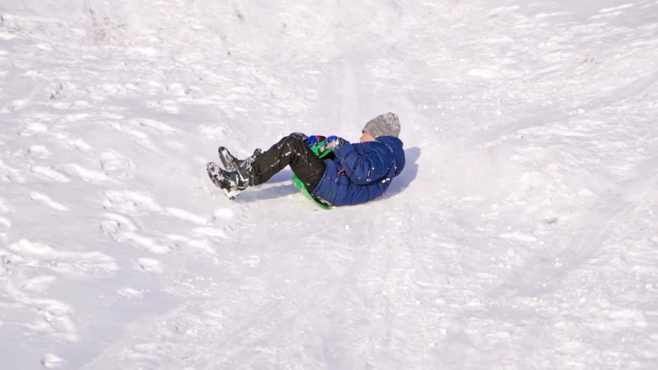 Boy sledding on a snow hill. Child riding down the slide on a plastic sled in winter. Happy childhood. Slow motion.