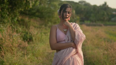 Mujer-Elegante-Con-Vestido-Rosa-Posando-Pensativamente-En-Un-Entorno-Rural,-Hora-Dorada