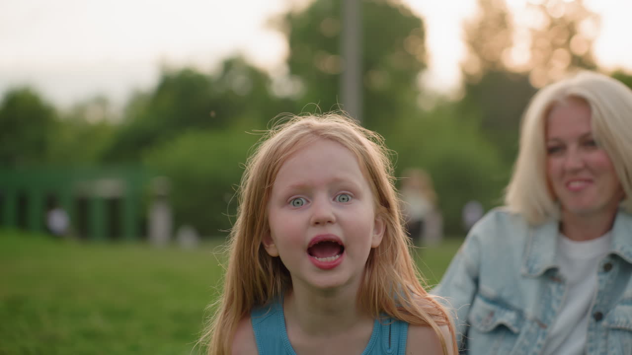 Bambina con gli occhi spalancati che parla giocosamente mentre la madre sorridente osserva dallo sfondo nel parco, alberi ed erba sfocati dietro, calda luce serale che cattura un momento familiare spontaneo.