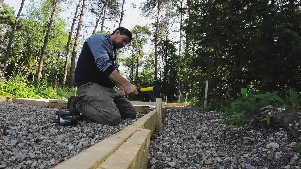 Carpenter Hitting Wood Stakes Into Ground With A Sledge Hammer. Formwork For Concrete Pad.