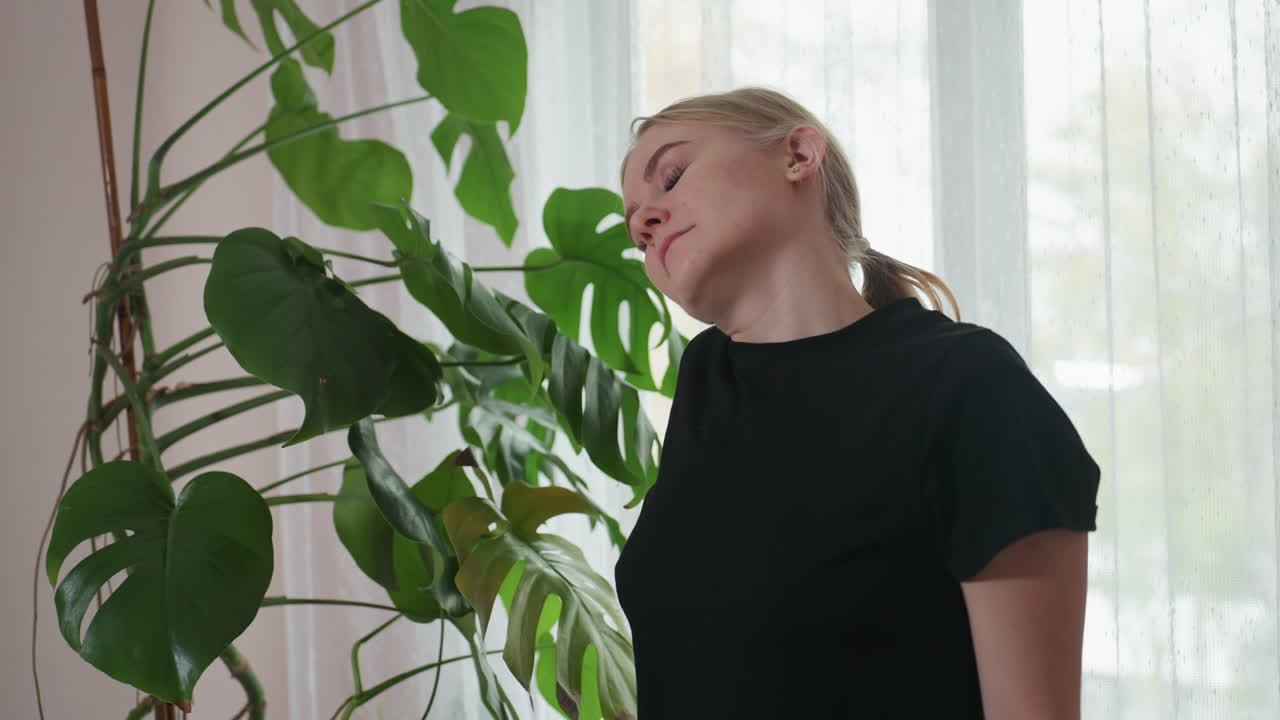 Massage therapist dressed in black gently stretching neck with closed eyes, seeking comfort and relaxation while standing beside vibrant indoor plant and sheer curtains in naturally lit room