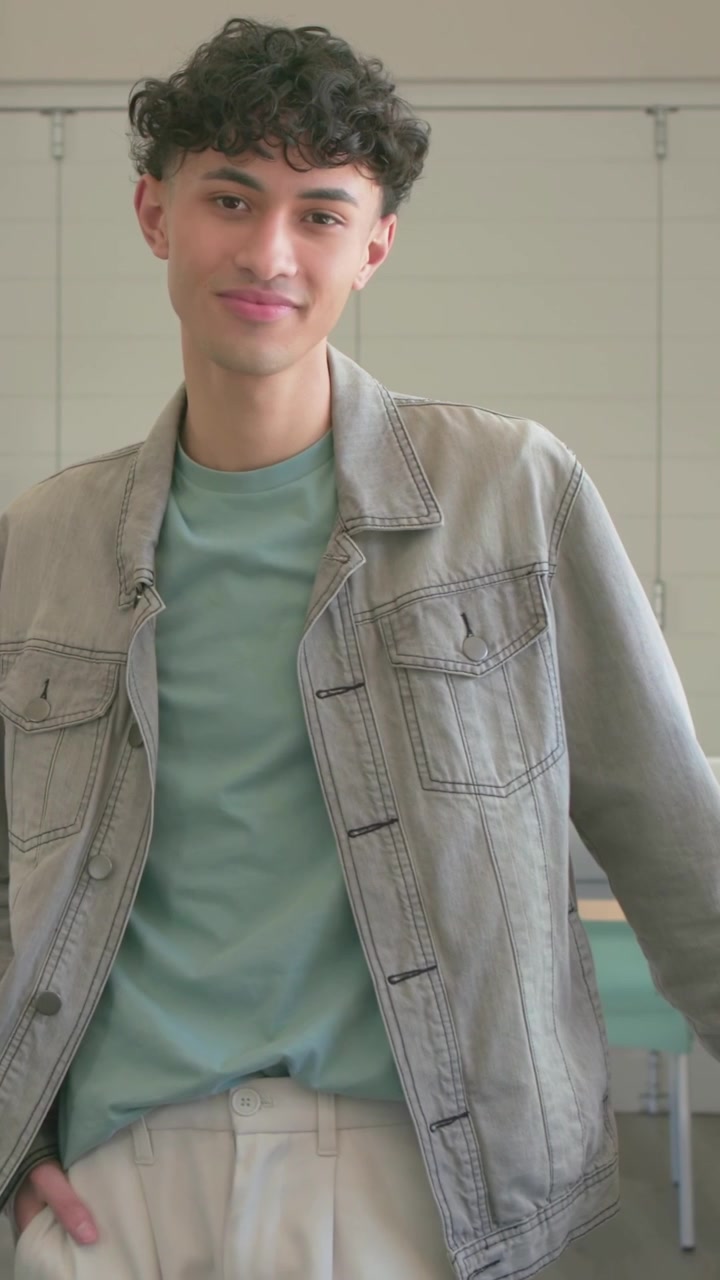 Vídeo vertical: Homem em pé se ajeitando, sorrindo em uma sala de aula, câmera gravando, microfone de lapela na jaqueta.