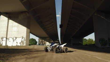 Goldenhour-dolly-push-in-shot-of-blue-and-white-atv-all-terrain-vehicle-quad-parked-on-dirt-under-a-bridge