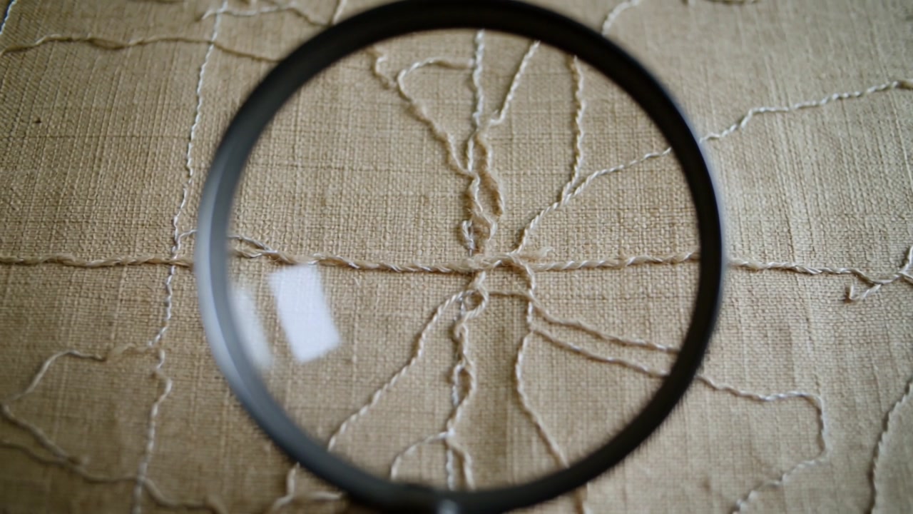 Sliding magnifying glass nudging down over tabletop textile, aligning stitched motif for exam