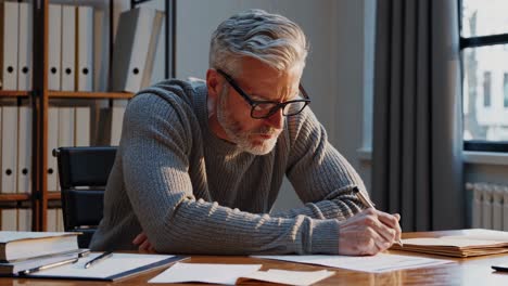 mature professional meticulously examines documents at office desk, showcasing concentration and attention to detail in a brightly lit workspace