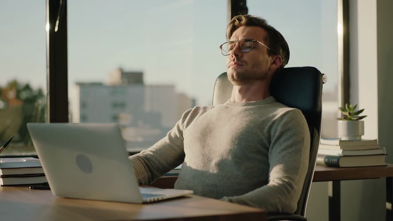 Appoggiato all'indietro, un uomo con un maglione leggero a maglia si ferma a pensare dopo essersi concentrato sul laptop alla scrivania.
