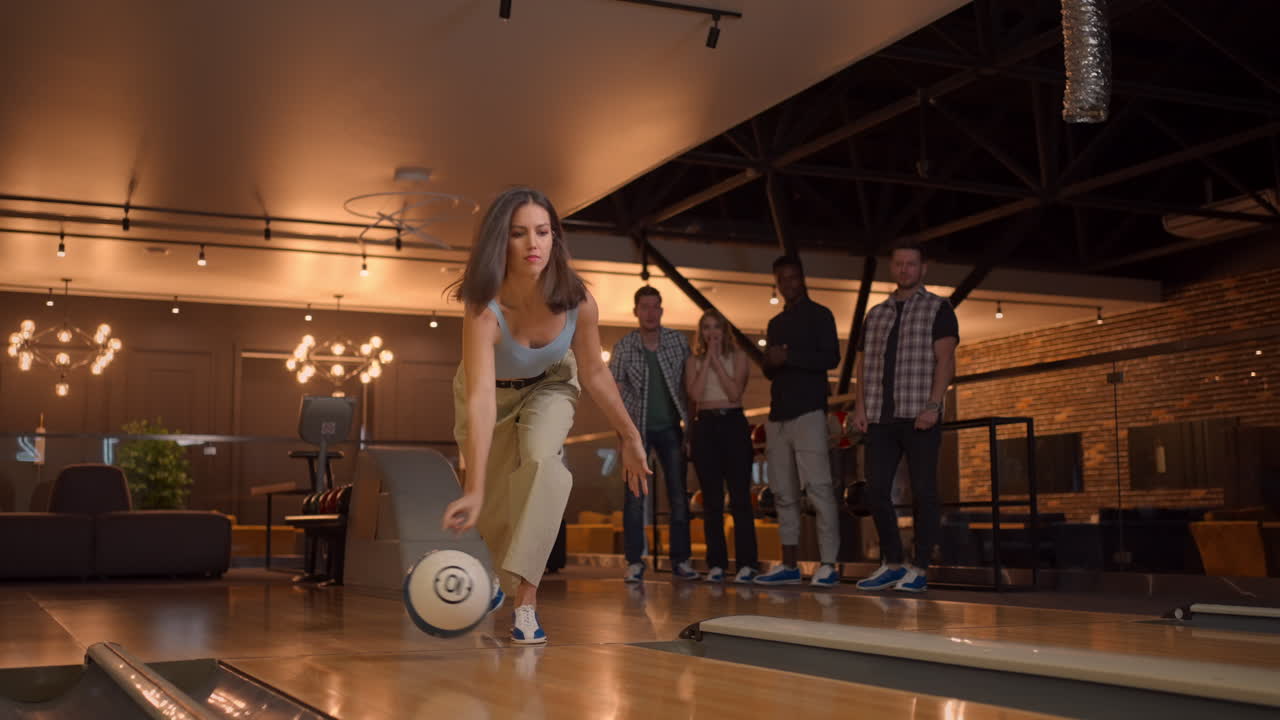 A woman in a bowling alley throws a ball on the track and knocks out a shot in slow motion and jumps and dances for joy. A group of multi-ethnic friends play bowling together.