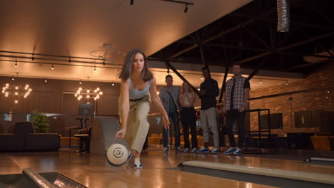 a woman in a bowling alley throws a ball on the track and knocks out a shot in slow motion and jumps and dances for joy. a group of multi-ethnic friends play bowling together.