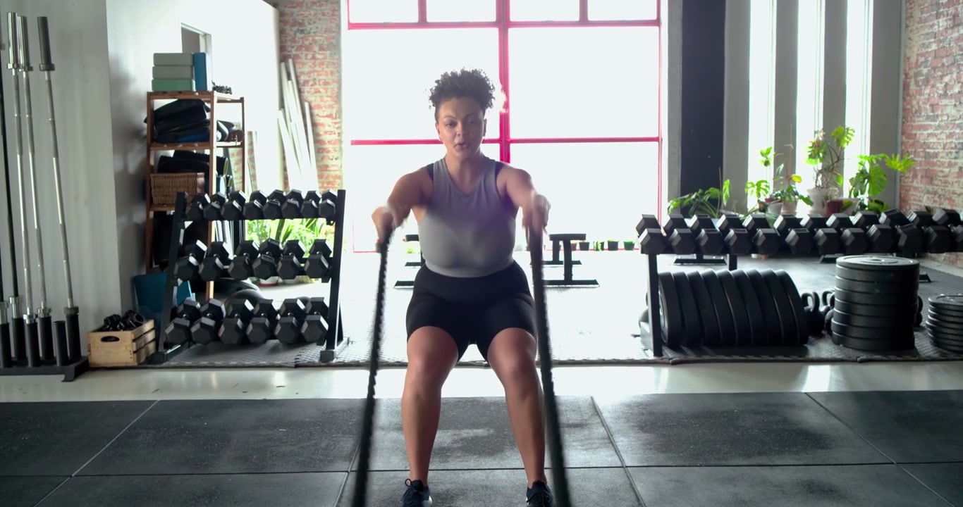 Donna afroamericana che termina le onde con la battle rope, tocca lo smartwatch monitorando la frequenza cardiaca in palestra
