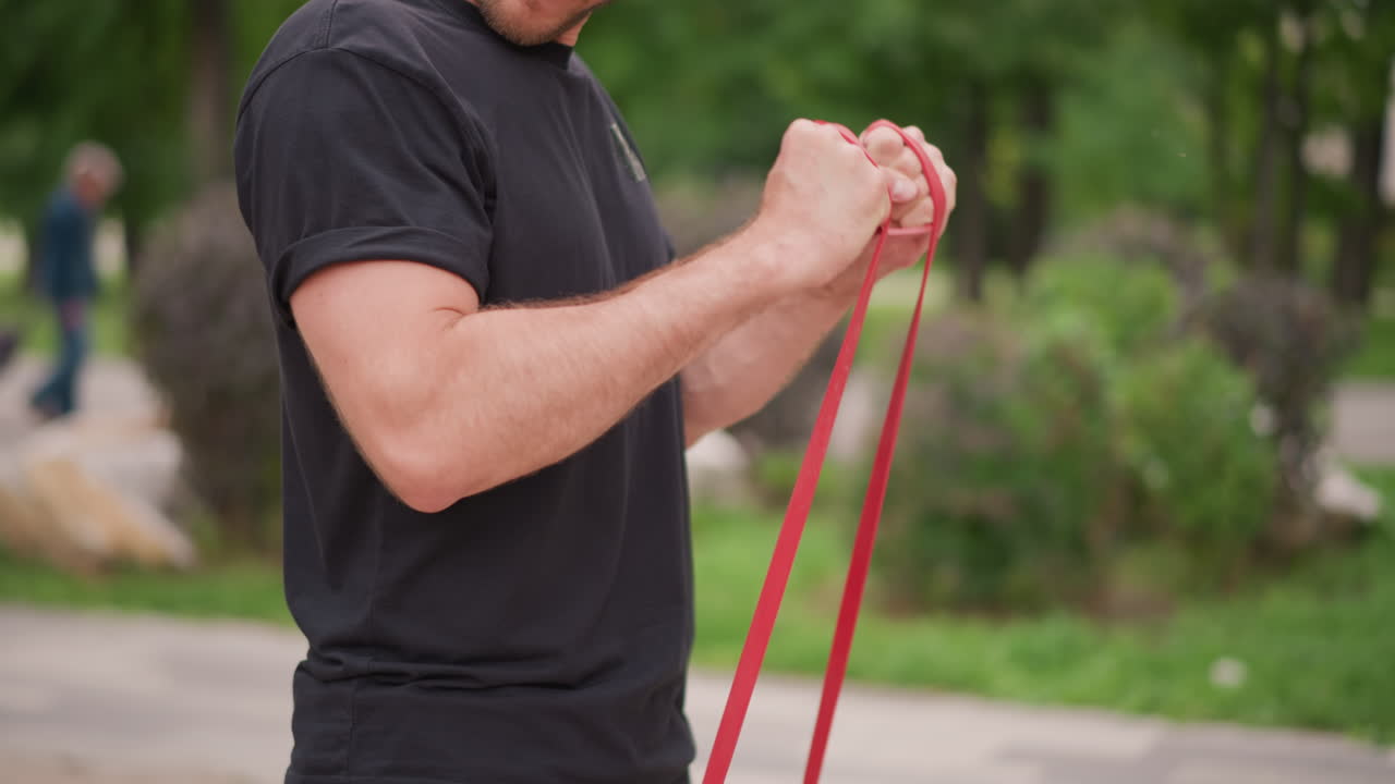 Man Exercising Shoulders, Person In Park Performing Shoulder Exercises With Resistance Band, Individual Practicing Controlled Shoulder Movements Using Resistance Band In Serene Outdoor Environment