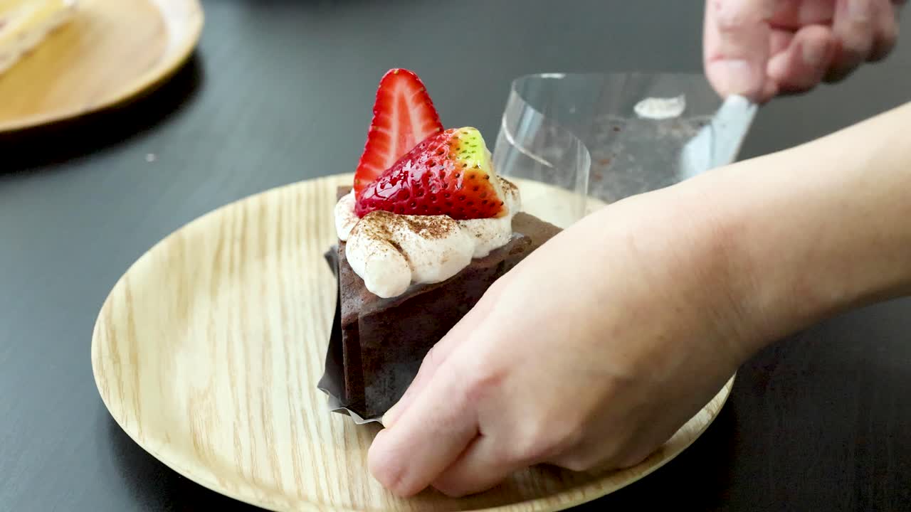 Hands carefully peel acetate from chocolate cake slice on wooden plate, natural soft lighting, close-up