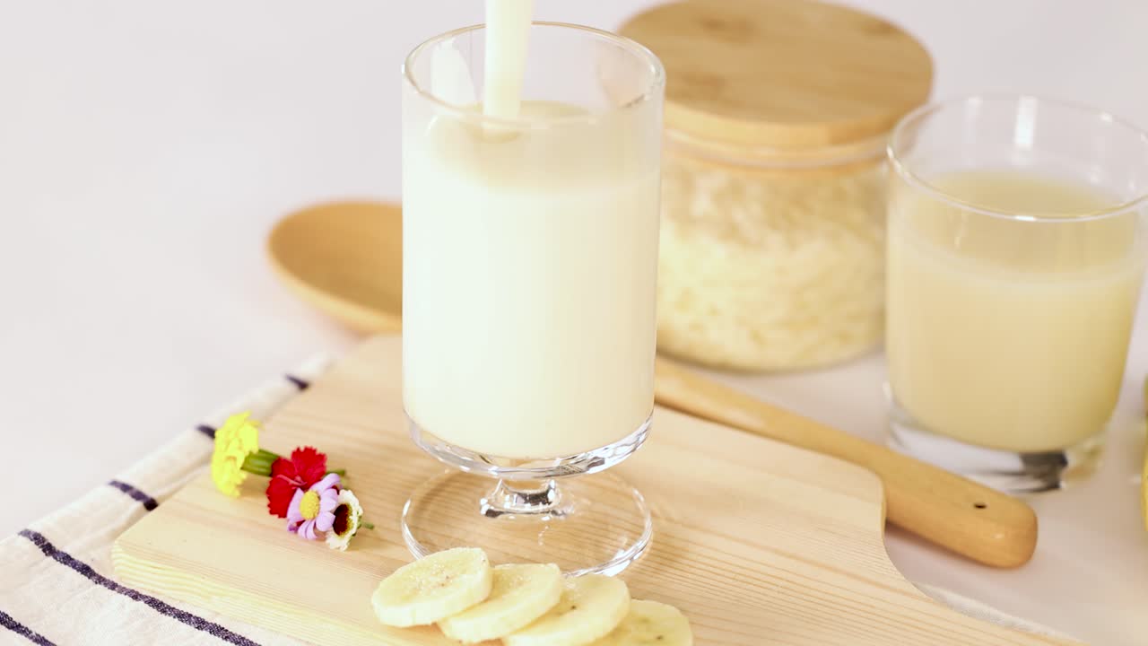 Banana milkshake poured into glass, surrounded by sliced banana, wooden spoon, and bright lighting