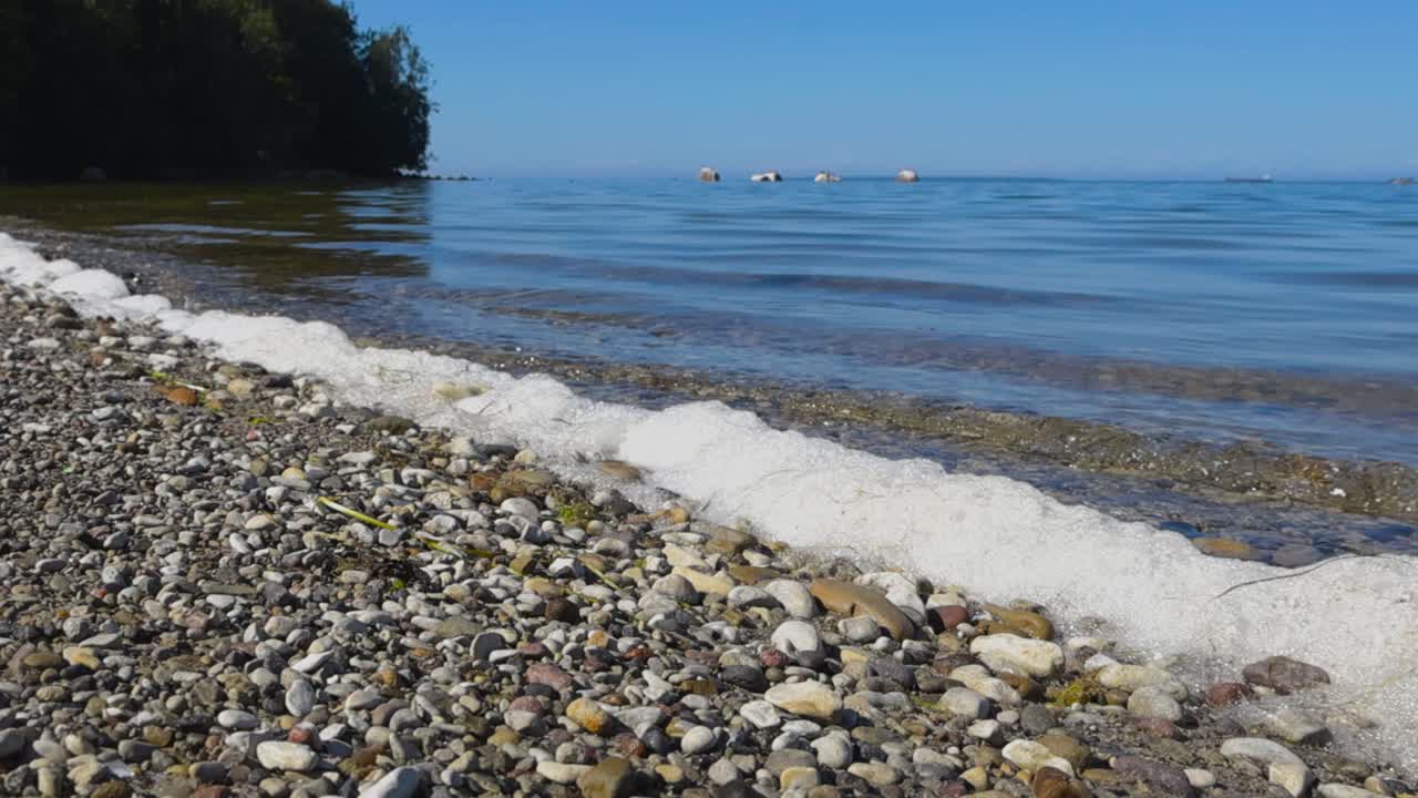 Low angle view of a rocky beach seaside shoreline with waves hitting the rocks and pebbles. White sea foam and bubbles visible that are struck by small waves during summer sunny day. Horizon visible