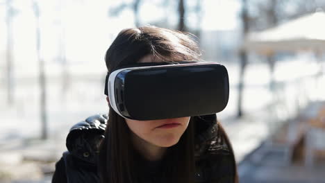 serious young dark-haired woman in warm black waistcoat wearing virtual reality glasses in early autumn park