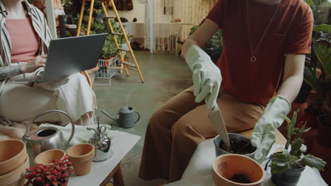 girl filling pot with soil and speaking with coworker in flower shop