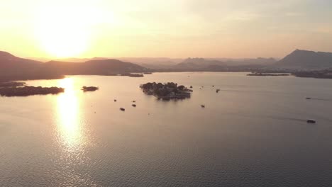 Vista-Dorada-Del-Lago-Pichola-Durante-La-Puesta-De-Sol-Con-El-Palacio-Del-Lago-Taj-En-El-Centro,-En-Udaipur,-Rajasthan,-India---Toma-Aérea-De-órbita-Amplia