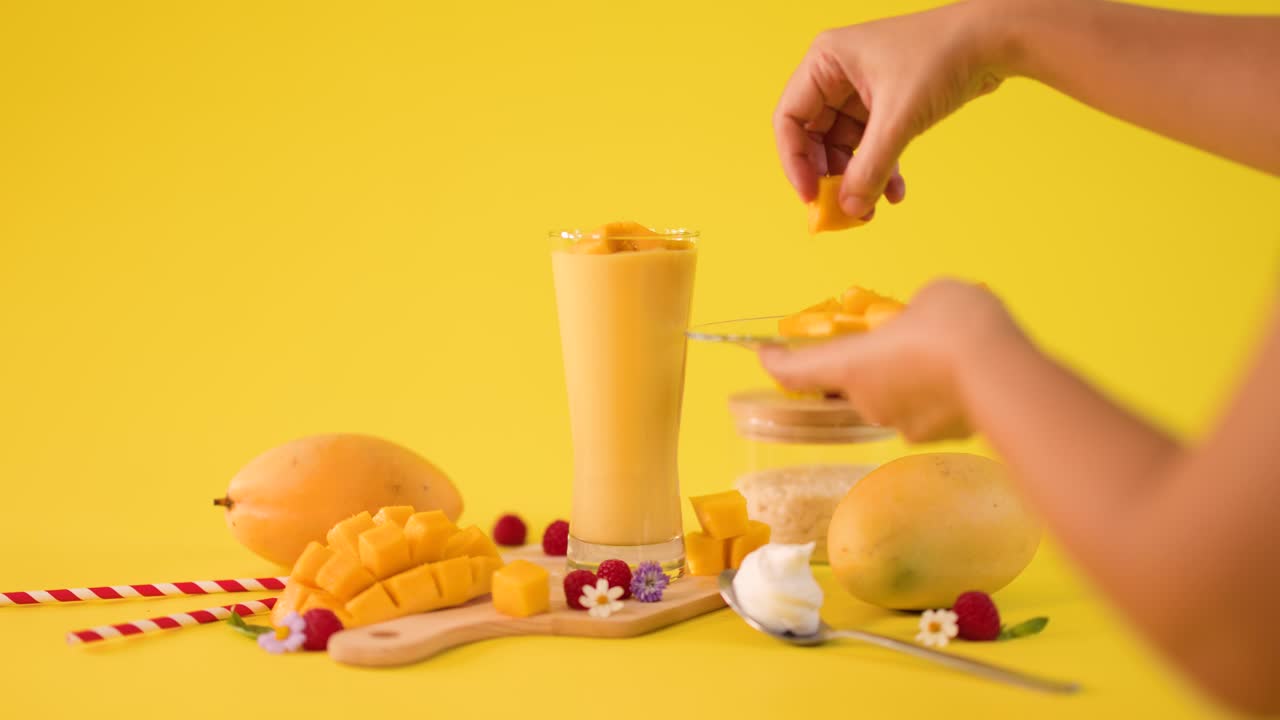 Mani umane decorano un frullato di mango alto accanto a frutta fresca su sfondo giallo brillante, telecamera statica