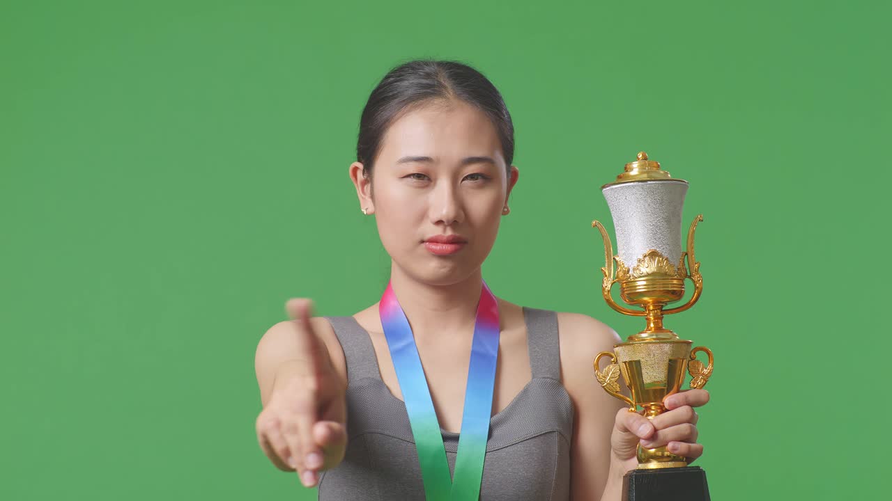 Close Up Of Asian Woman With A Gold Medal And Trophy Disapproving With No Index Finger Sign And Smiling To Camera On Green Screen Background In The Studio
