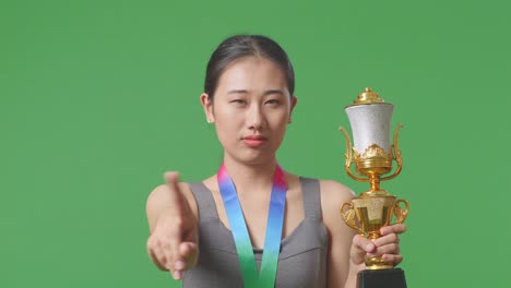 close up of asian woman with a gold medal and trophy disapproving with no index finger sign and smiling to camera on green screen background in the studio