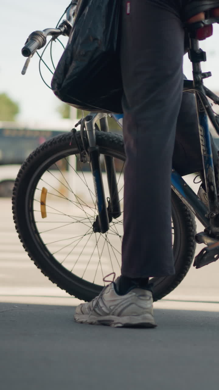 Urban cyclist waiting at crosswalk with bicycle wheel and sneaker visible, carrying black tote bag, standing on asphalt under soft morning light, closeup of tire, spokes and handlebar, casual commuter