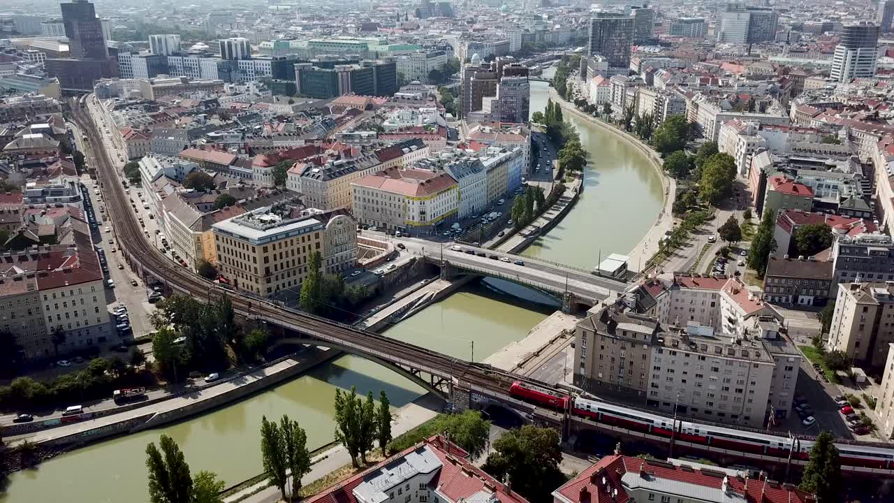 Train on bridge crossing Danube River in Vienna during summer season. Aerial tilt up wide shot. Modern apartment housing area in suburb. Colorful trees in autumn season. Downtown in background.